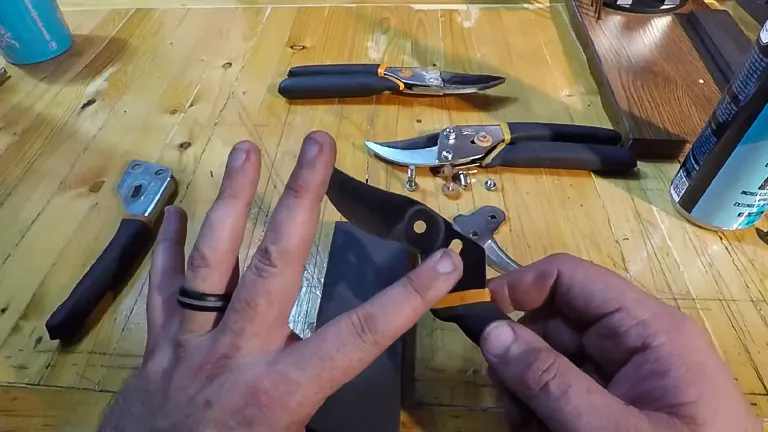 A person's hands assembling or sharpening Fiskars Pruning Shears on a wooden workbench with various disassembled shear parts and a can of lubricant visible.