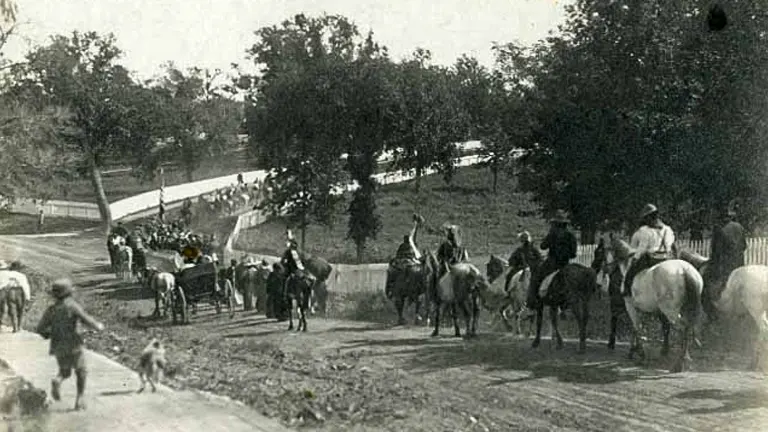 Vintage black and white photo of people on horseback and a few on foot on a dirt road, with trees in the background, suggesting a community or social event from an earlier era.
