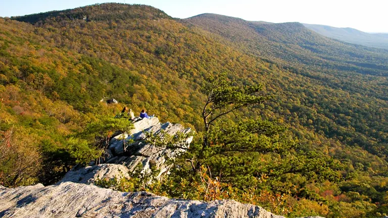 People sitting on rocky outcrops enjoying the view of a forested mountain range in the early fall, with leaves starting to change color under the soft glow of the setting sun.