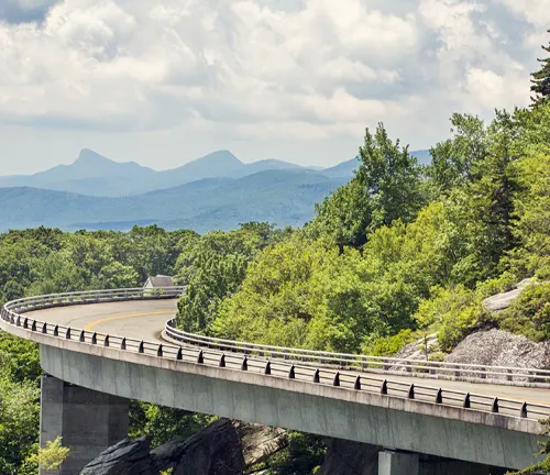 A scenic view of the Linn Cove Viaduct curving around the lush greenery of the Blue Ridge Mountains on a sunny day with fluffy clouds in the sky.