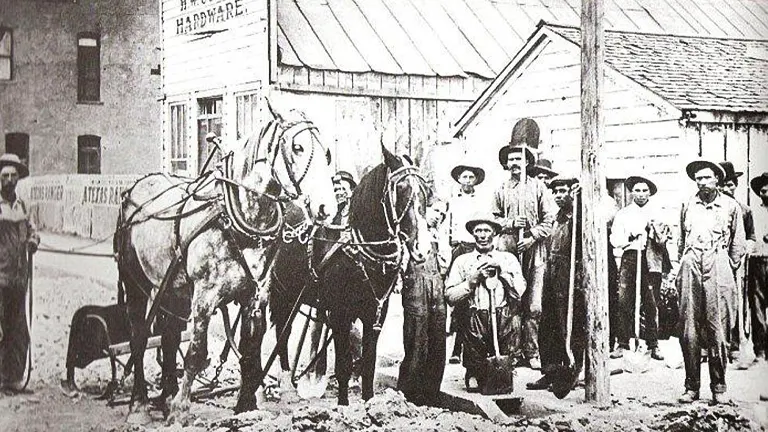 Historical black and white photo showing a group of people and a team of horses in front of "Hughes Hardware" store, with individuals wearing period clothing and hats indicative of late 19th or early 20th century American frontier life.