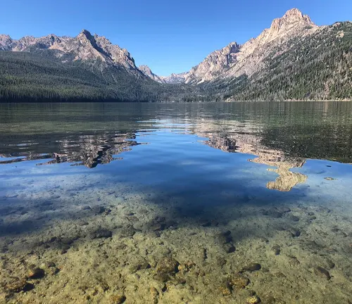 Clear, calm waters of a mountain lake reflecting rugged peaks under a blue sky, with the lakebed visible through the transparent water.