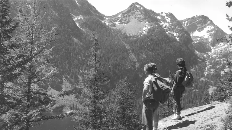 Black and white historical photo of two hikers with backpacks standing on a rocky overlook, gazing at a mountainous landscape with dense forests and a hidden lake below, evoking a sense of exploration and adventure.