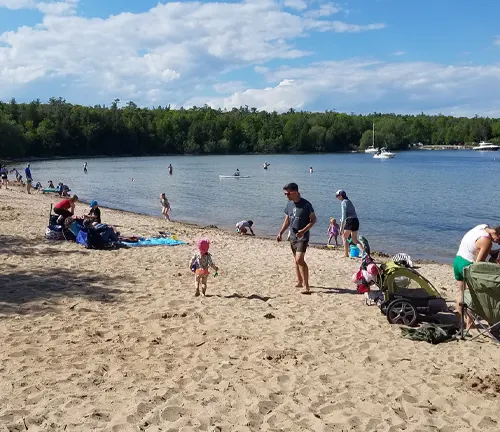Families and visitors enjoy a sunny day on a sandy beach at Peninsula State Park, with swimmers in the water and boats anchored near the tree-lined shore.