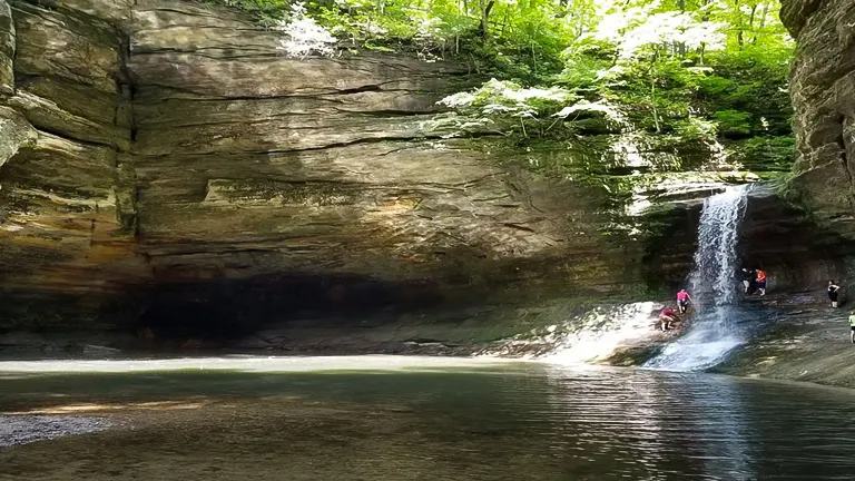 Image of people enjoying a natural waterfall and pool at the base of a shaded rock cliff, with sunlight filtering through the surrounding foliage above.