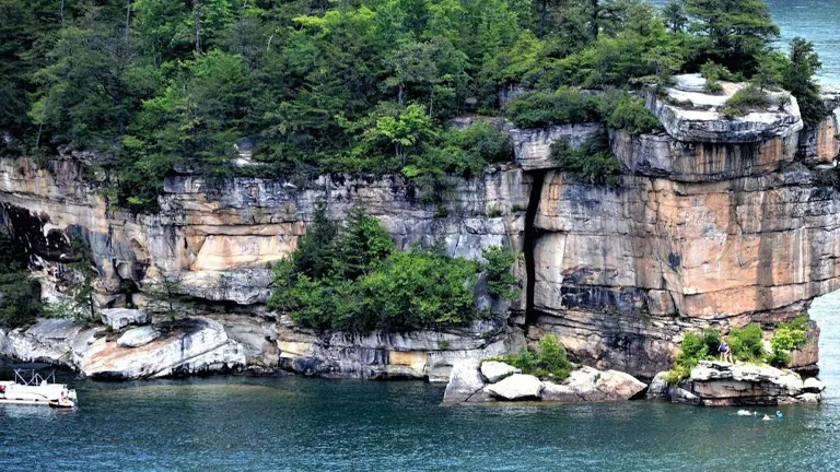 Close-up view of a majestic, stratified rock cliff adorned with greenery, overhanging the clear waters of a lake with a small boat floating nearby and people visible on a rocky outcrop.
