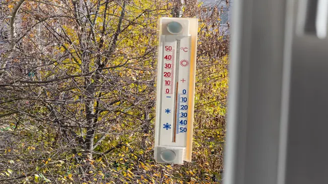 Thermometer showing temperature in Celsius attached to a window with autumn trees in the background