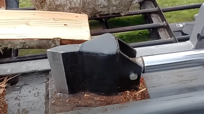 Close-up of the wedge end of a log splitter with wood chips around it and split logs in the background.