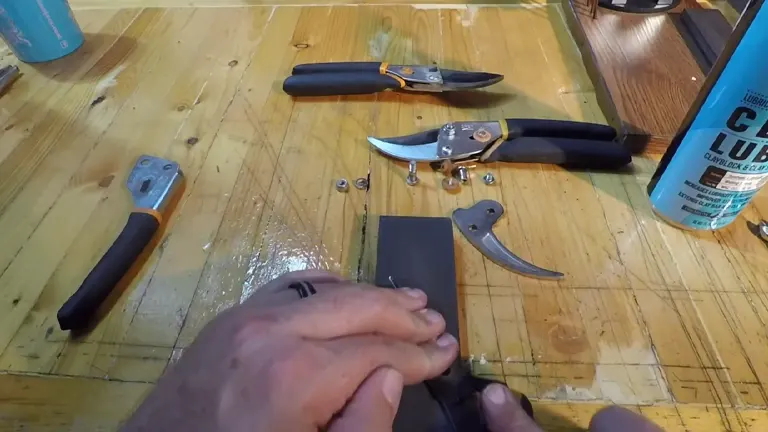 Hands sharpening a blade of Fiskars Pruning Shears with a sharpening stone on a wooden workbench, surrounded by various shear components and a can of lubricant.
