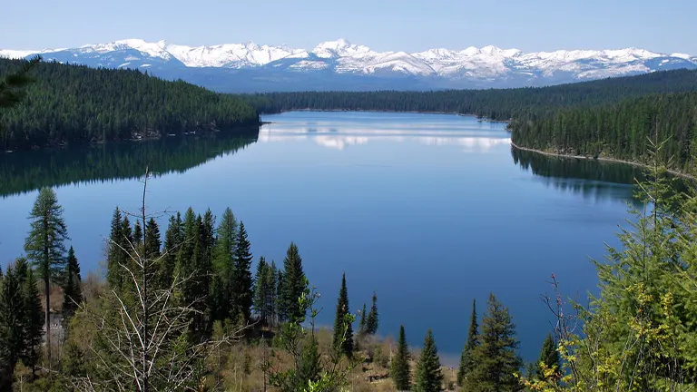 Calm blue lake surrounded by coniferous forest with snow-capped mountain range in the background under a clear sky, reflecting a tranquil and scenic landscape.
