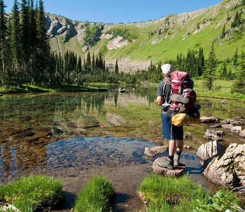 A hiker with a large backpack stands on rocks at the edge of a clear mountain lake, surrounded by green pine trees and rolling hills, reflecting on the peaceful water in front of them.