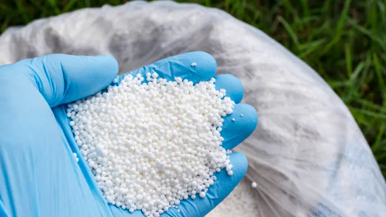 A gloved hand holding a handful of white granular synthetic fertilizer with a large sack in the background, ready for application in agriculture or gardening.