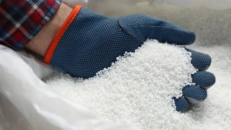 Close-up of a gloved hand holding white granular material, possibly fertilizer, above a large bag.