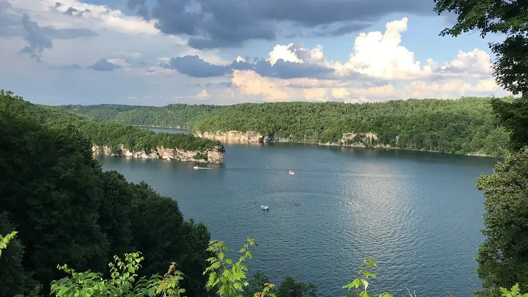 A serene lake view surrounded by dense forests with a dramatic cliff face in the distance, under a sky with puffy clouds, with boats gently floating on the water's shimmering surface.
