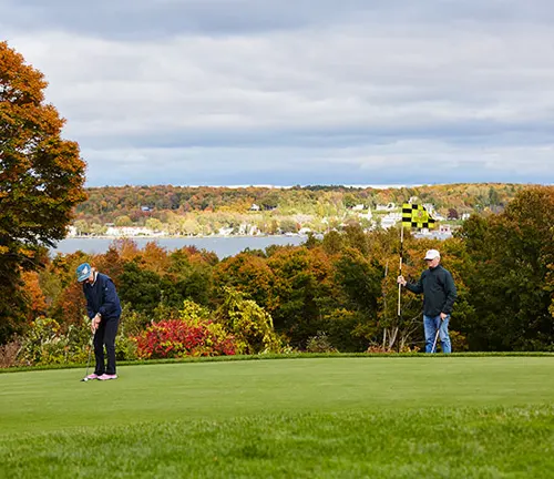 Two people are playing golf on a lush course with vibrant autumn trees in the background, overlooking a scenic view of a distant town by the water.