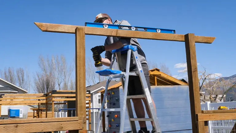 A person on a ladder using a level on top of a wooden garden fence archway, with clear blue skies and mountainous background, indicating precision work during a sunny day.
