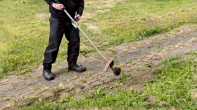 A person in black is holding a long-handled garden tool with a curved metal blade over mixed grass and paving stones.