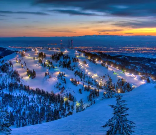 Twilight over a snow-covered ski resort with lit ski runs snaking through pine trees, overlooking a distant cityscape illuminated against the backdrop of a dramatic sunset and mountain silhouette.