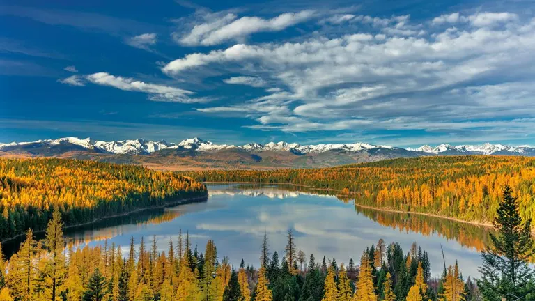 Scenic view of a tranquil lake reflecting the sky, surrounded by a forest with autumn-colored foliage, and a majestic mountain range with snow-capped peaks in the background under a vast blue sky with streaky clouds.