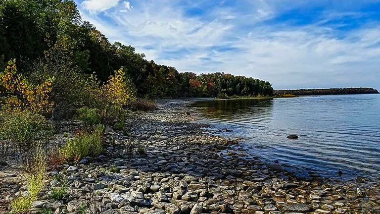 A tranquil lakeside view with a rocky shore in the foreground, transitioning to colorful autumn trees and calm waters, under a sky with high clouds.