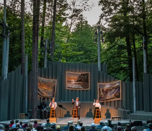 An outdoor theater performance in progress, with actors on stage and an audience in a natural amphitheater surrounded by tall trees.