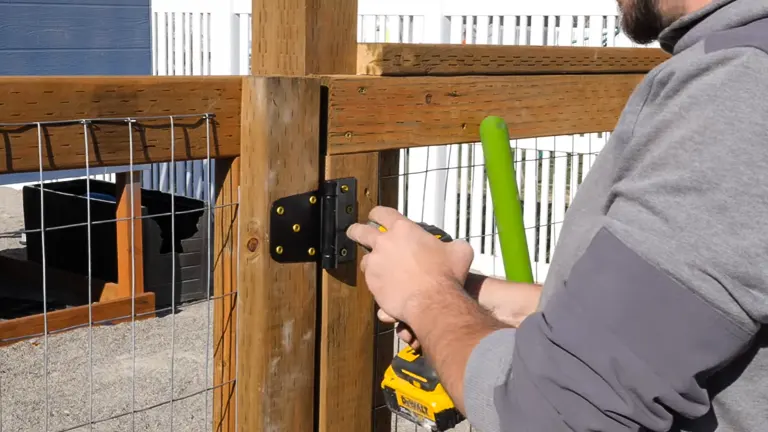 Close-up of a person's hands installing a black metal hinge onto a wooden gate post with a drill, part of a garden fence assembly process.