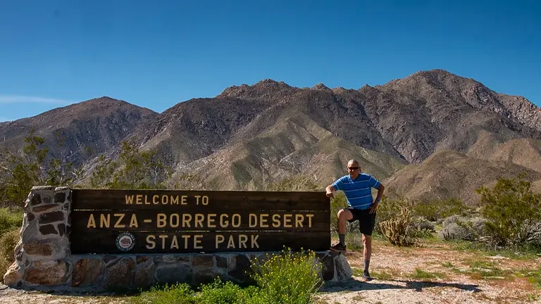 A man posing next to the welcome sign of Anza-Borrego Desert State Park with mountainous terrain in the background.
