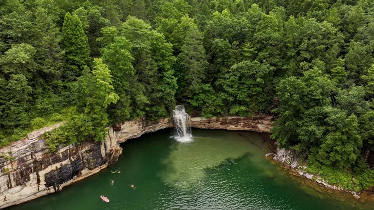 Aerial view of a secluded waterfall cascading into a green, tranquil lake surrounded by dense forests. A few people and a raft are visible, enjoying the water near the waterfall's base