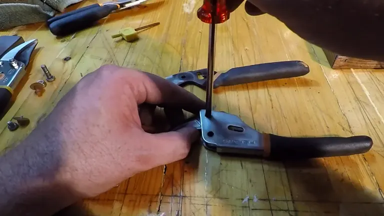 A person's hands are using a screwdriver to reassemble the handle and blade of Fiskars Pruning Shears on a wooden workbench, with parts and tools laid out around them.