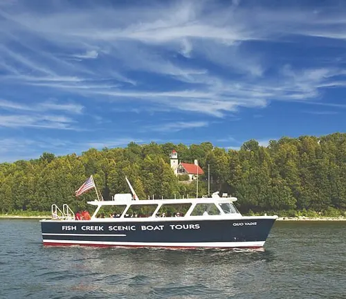 The 'Fish Creek Scenic Boat Tours' vessel glides on clear blue waters with the Eagle Bluff Lighthouse and lush green trees in the background, under a bright blue sky.