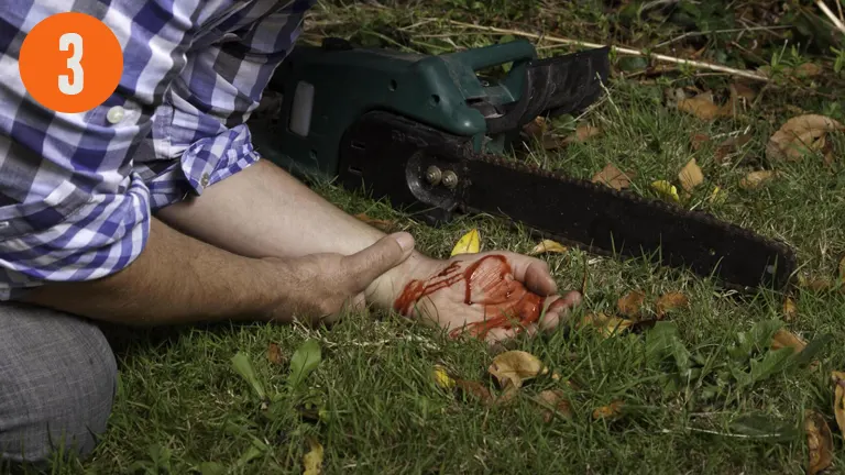 person in a checkered shirt sitting on the grass next to a chainsaw possibly injured