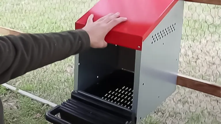 Hand placing a red and grey chicken nesting box inside a coop with chicken wire