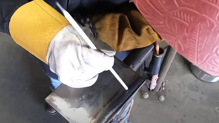 Welder's gloved hands holding a stick electrode over a metal workpiece with a welding bead