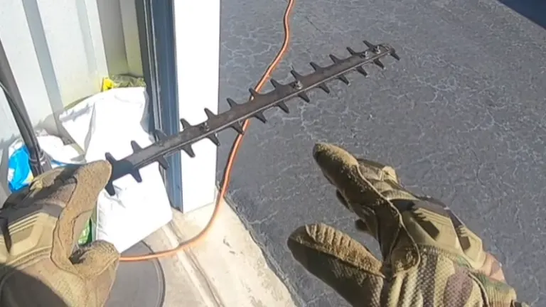 Hand in camouflaged glove holding a black hedge trimmer with an orange cord near a building.