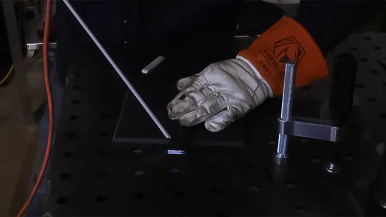 Hand in a welding glove placing a welding rod on a metal workpiece on a clamped welding table