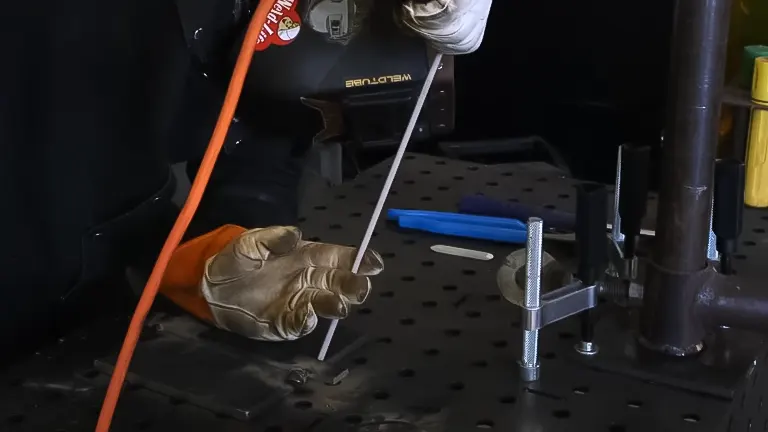 A welder's gloved hand holding a 7018 welding electrode over a metal workpiece on a welding table