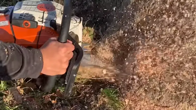 Person using a chainsaw to cut through a tree stump or log, with wood chips flying away from the cutting area.