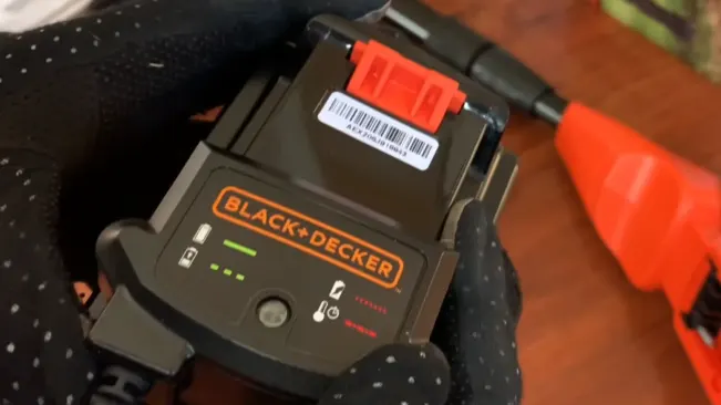 Person in gloves holding a BLACK+DECKER battery pack with charge level indicator lights, with a glimpse of an orange tool in the background