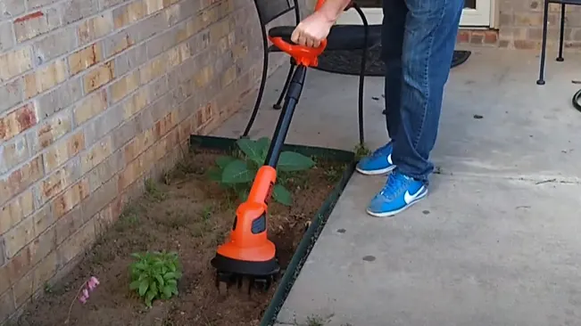Person using an orange trimmer on weeds near a brick wall