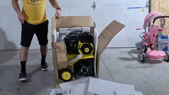 person, wearing black shorts and a yellow t-shirt, unboxing a new yellow and black machine in a garage