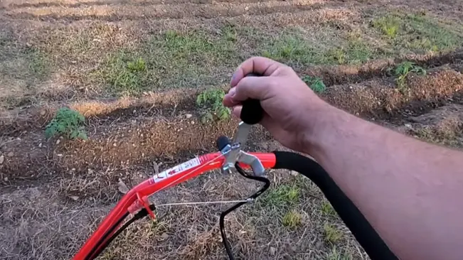 hand gripping the handlebar of a red tiller, with a cultivated garden in the background.