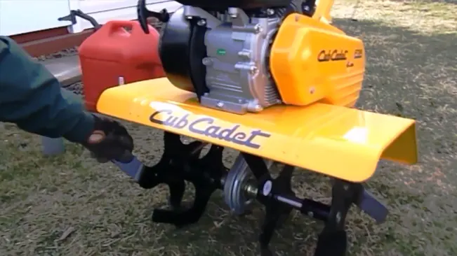 a person adjusting a bright yellow Cub Cadet garden tiller with black metal tines, set against a background of grass and a red gasoline container