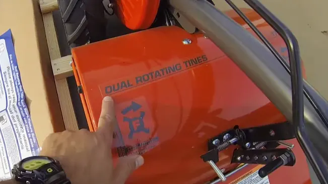 person’s hand pointing at the label “DUAL ROTATING TINES” on an orange mechanical device, surrounded by various mechanical parts
