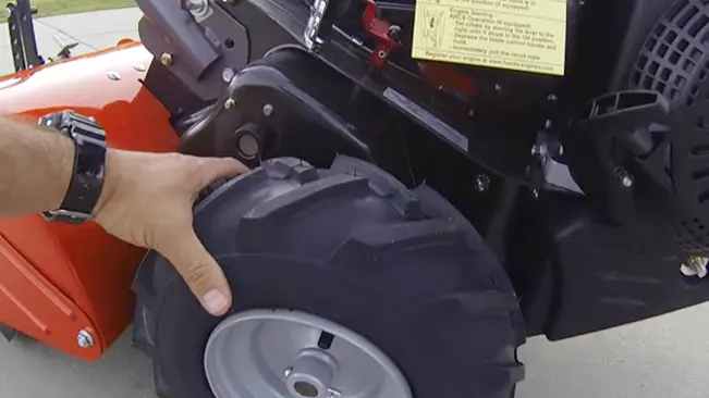 person’s hand touching the wheel of an orange mechanical device, possibly a lawnmower or small tractor, with a visible warning label