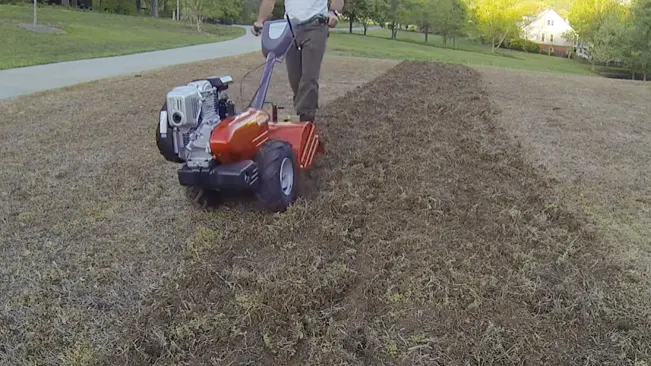 a person operating a motorized tiller on a patch of freshly tilled land, with greenery in the background