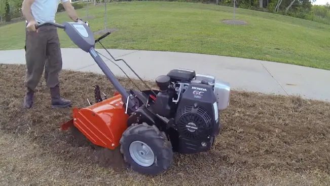 a person operating a red and black Honda-powered tiller on a grassy field, adjacent to a concrete pathway. The field appears recently tilled with upturned soil visible