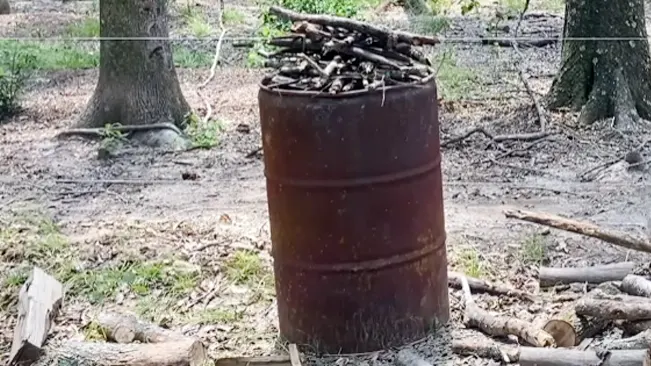 a close-up of a rusty, old barrel filled with sticks and twigs, standing in a wooded area with trees in the background