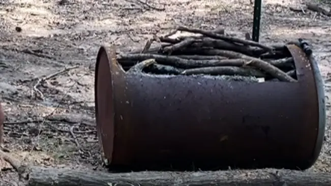 Rusted barrel full of logs on dirt field.