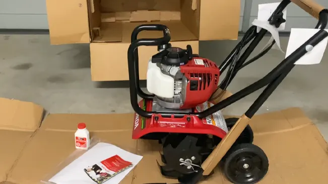 new red tiller on its cardboard packaging in a storage area, with an instruction manual and a bottle of engine oil nearby