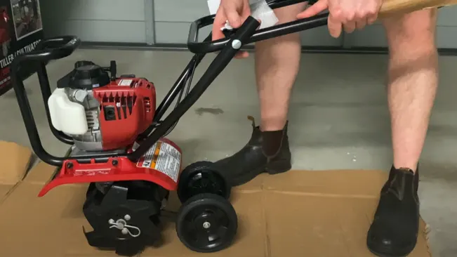 a person operating a new red rototiller with black wheels and handles on a cardboard surface indoors